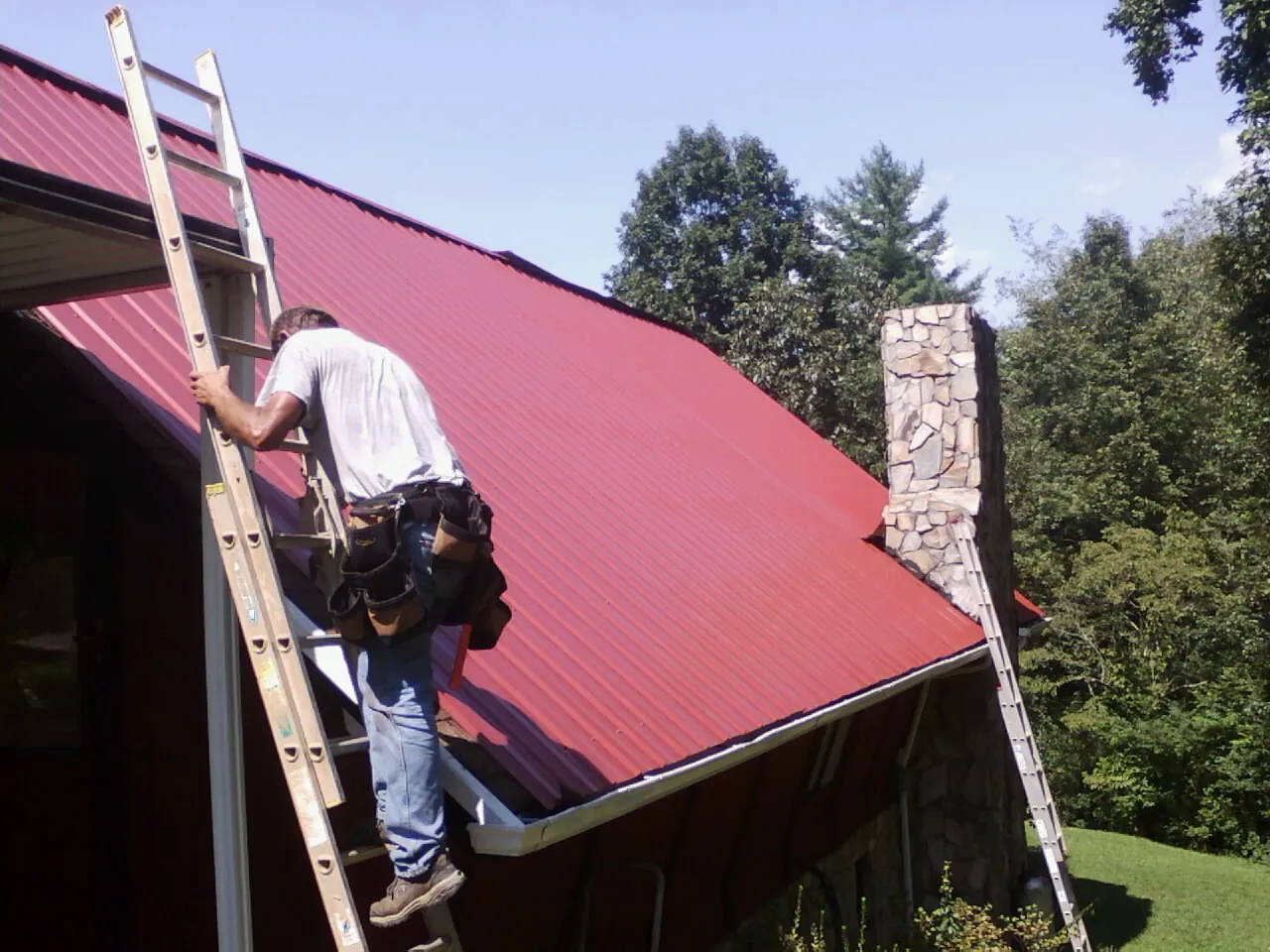 Roofer on a ladder beside a red metal roof and stone chimney — Soffit & Fascia Repair by Roofing Brothers in Ardsley